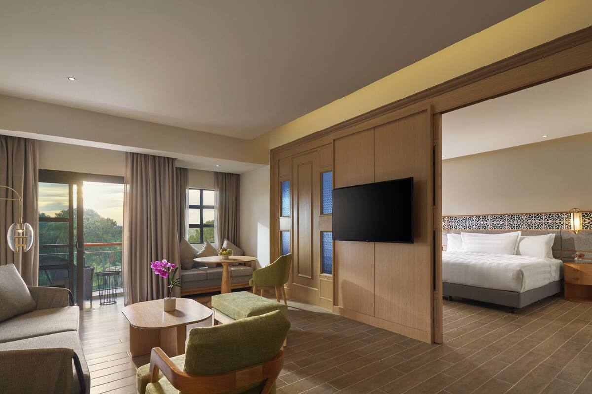 Een hotelkamer met een bank, fauteuil, klein tafeltje en een bed. Er is een televisie aan de muur bevestigd en een raam met gordijnen.