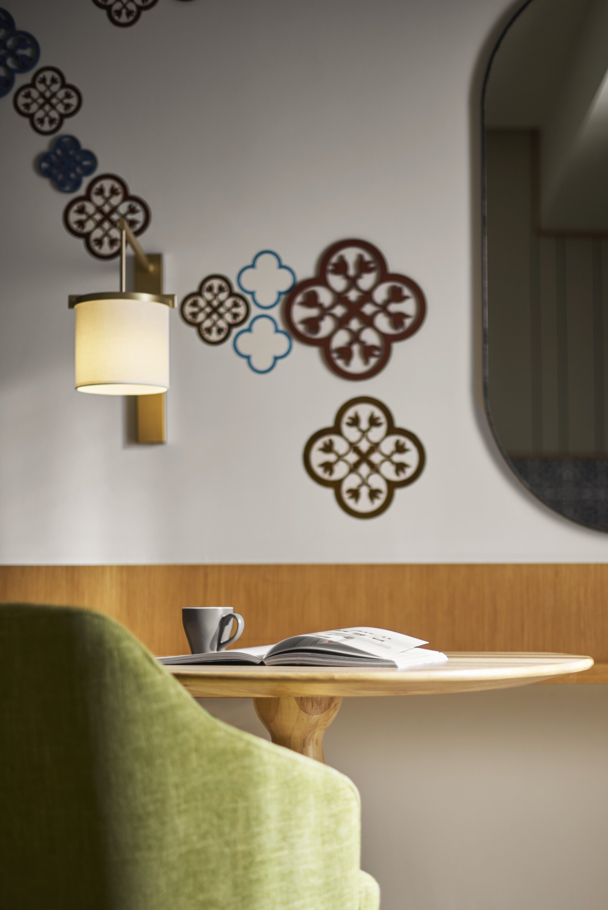 A wall with decorative patterns, a lamp, a table with a book and a cup, and a green chair.