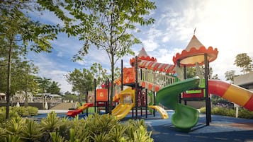 Explore the playground surrounded by lush greenery and trees, featuring slides and climbing elements on a safe rubberized surface.
