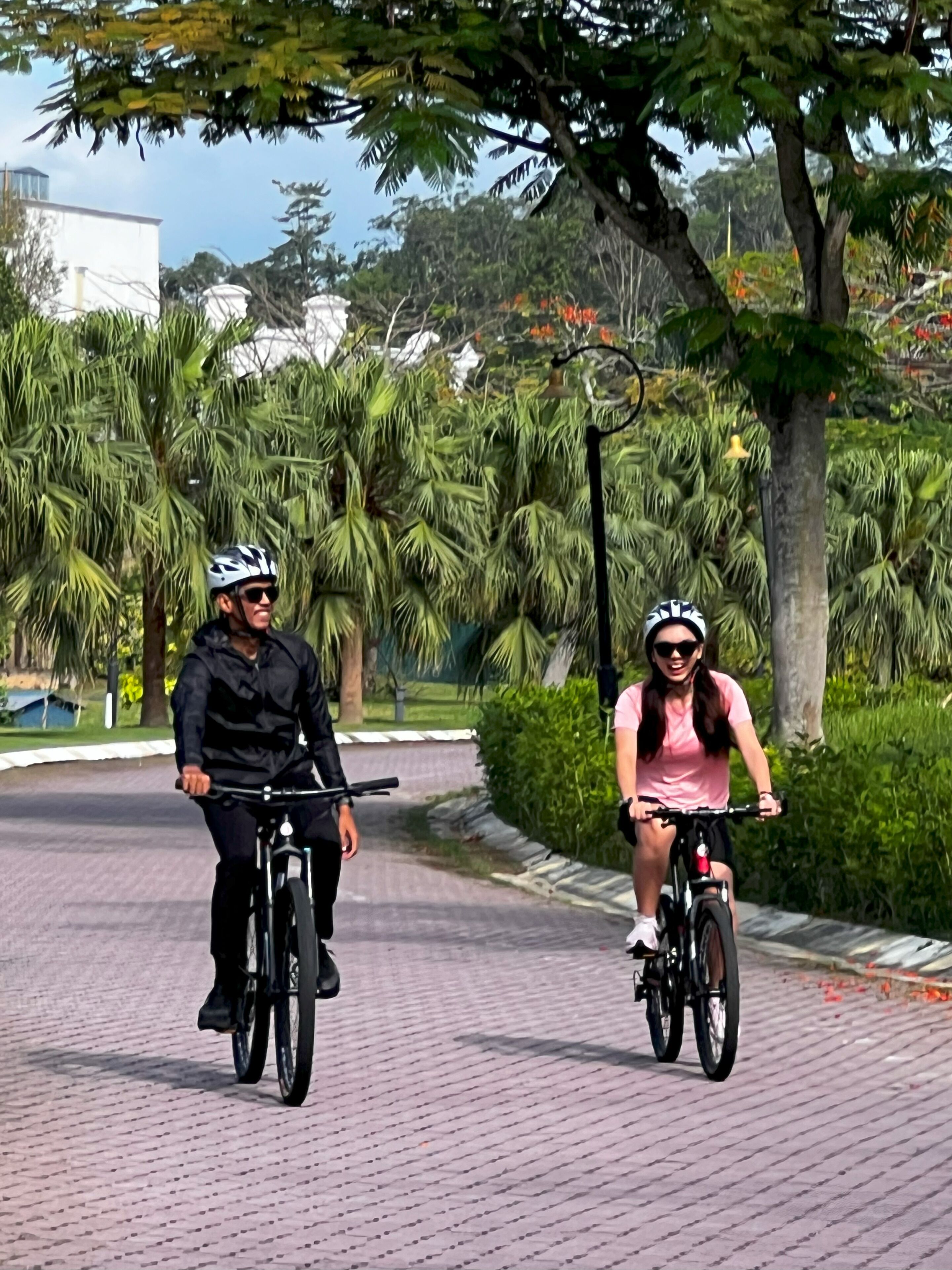 Two people riding bicycles on a paved path surrounded by greenery.