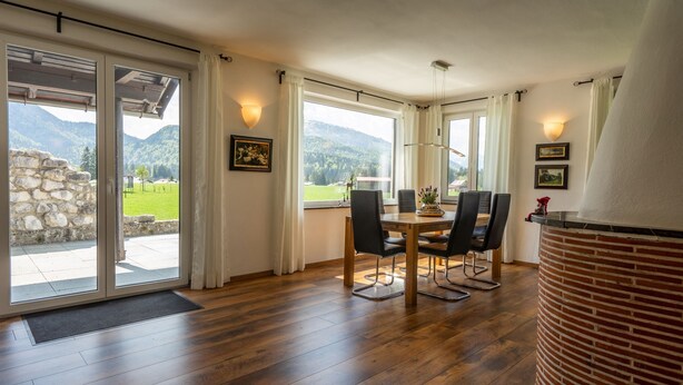 Ein Essbereich mit Tisch und Stühlen, großen Fenstern mit Vorhängen und Blick auf Berge.