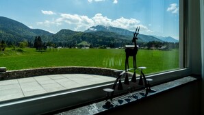Blick auf einen See mit Bergen im Hintergrund, sichtbar durch ein Fenster, eine Hirschskulptur und ein kleiner Tisch mit einer Lampe.