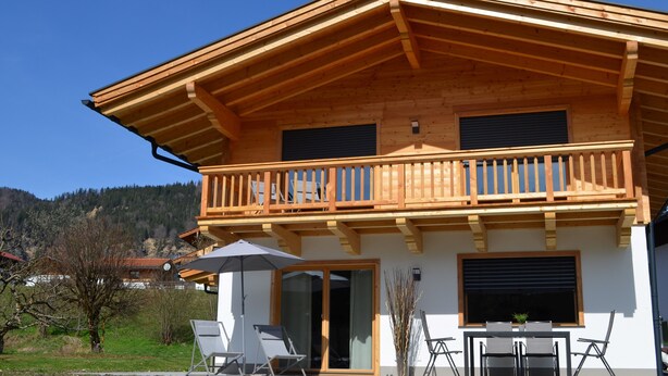 A two-story wooden house with a balcony, a sliding glass door, and a patio area with chairs and an umbrella. - Chalet 125qm für Max. 6 Personen, 3 Sep. Schlafzimmer (Reit im Winkl)