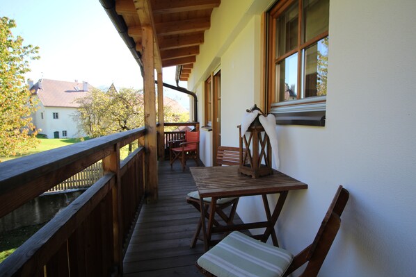 A wooden deck with a table and chairs, a window with a view of a house and trees, and a railing. - Im Winkl - "waldblick" / Gemütliche Ferienwohnung im 2. Obergeschoss (Dietenheim)