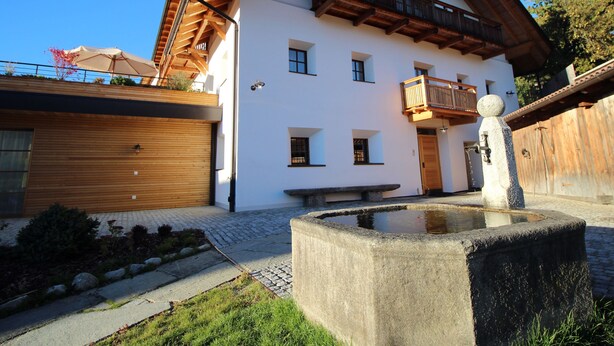 Een gebouw met twee verdiepingen, een houten balkon, een stenen fontein en een houten garagepoort.