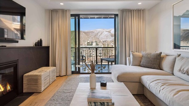 A modern living room with a fireplace, a sofa, a coffee table with a book, and a view of a mountainous landscape through large windows.