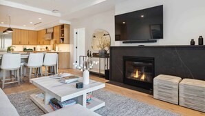 A modern living room with a fireplace, a flat-screen TV mounted above it, and a kitchen area with wooden cabinets and white bar stools.