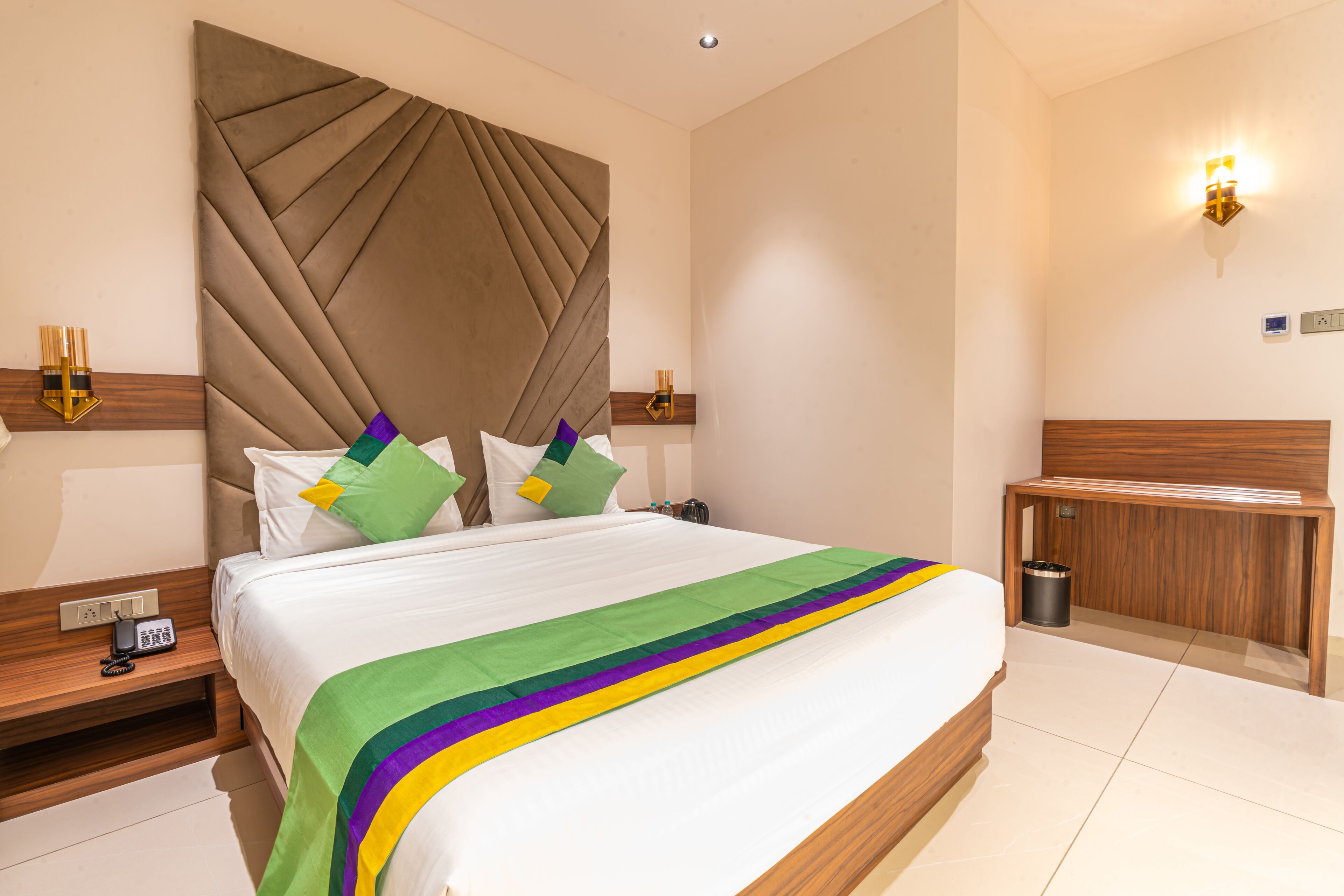 A hotel room with a large bed, a wooden headboard, bedside tables with lamps, a small wooden cabinet, and a trash can.