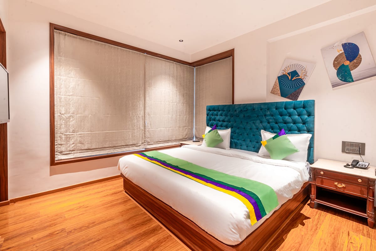 A bedroom with a large bed, wooden floor, a window with blinds, a nightstand, and artwork on the wall.