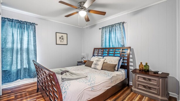 Un dormitorio con cama de madera, mesita de noche, cómoda, ventilador de techo y cortinas azules.