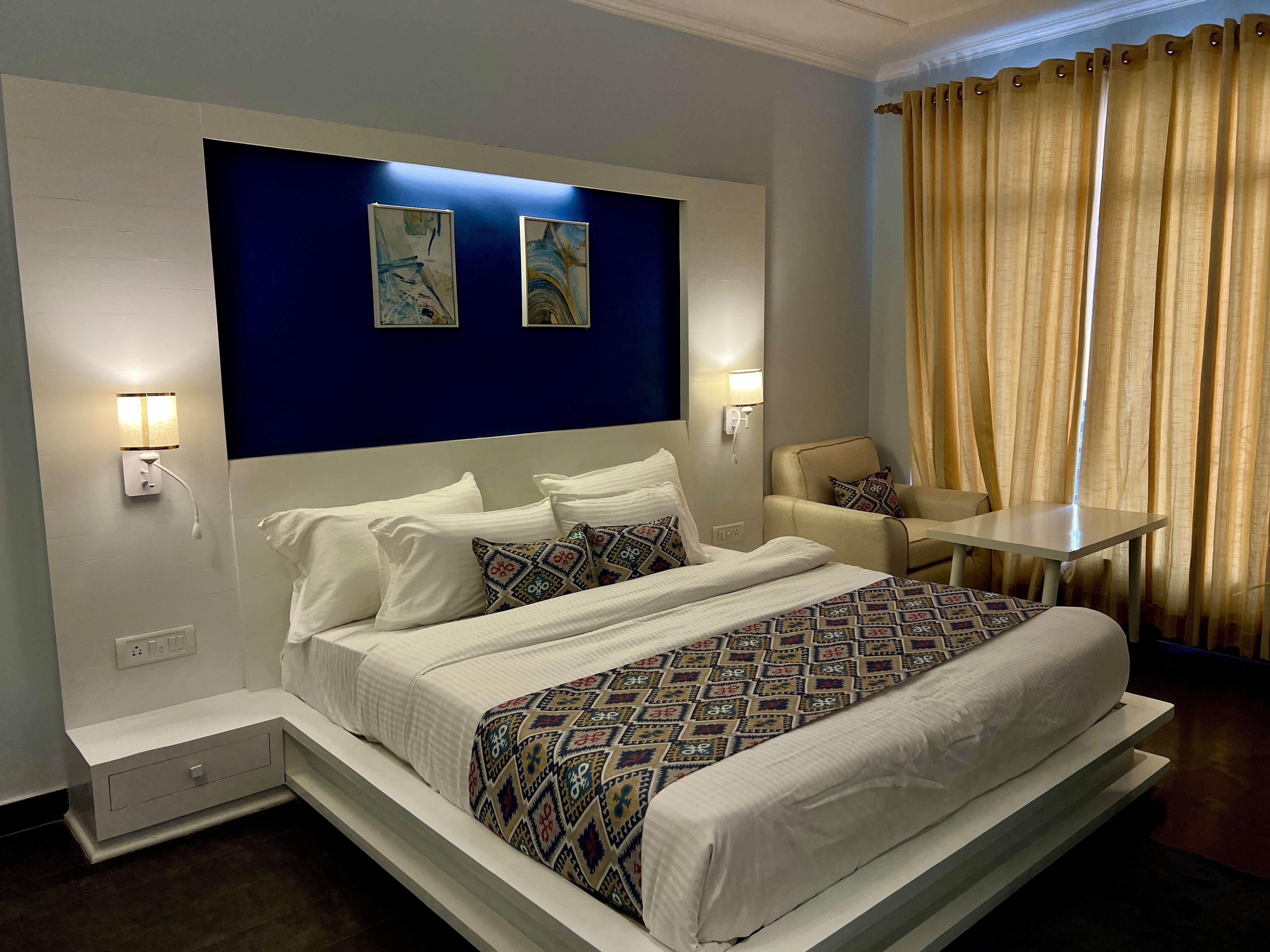 A hotel room with a large bed, two wall-mounted lights, a small table, a chair, and a window with curtains.