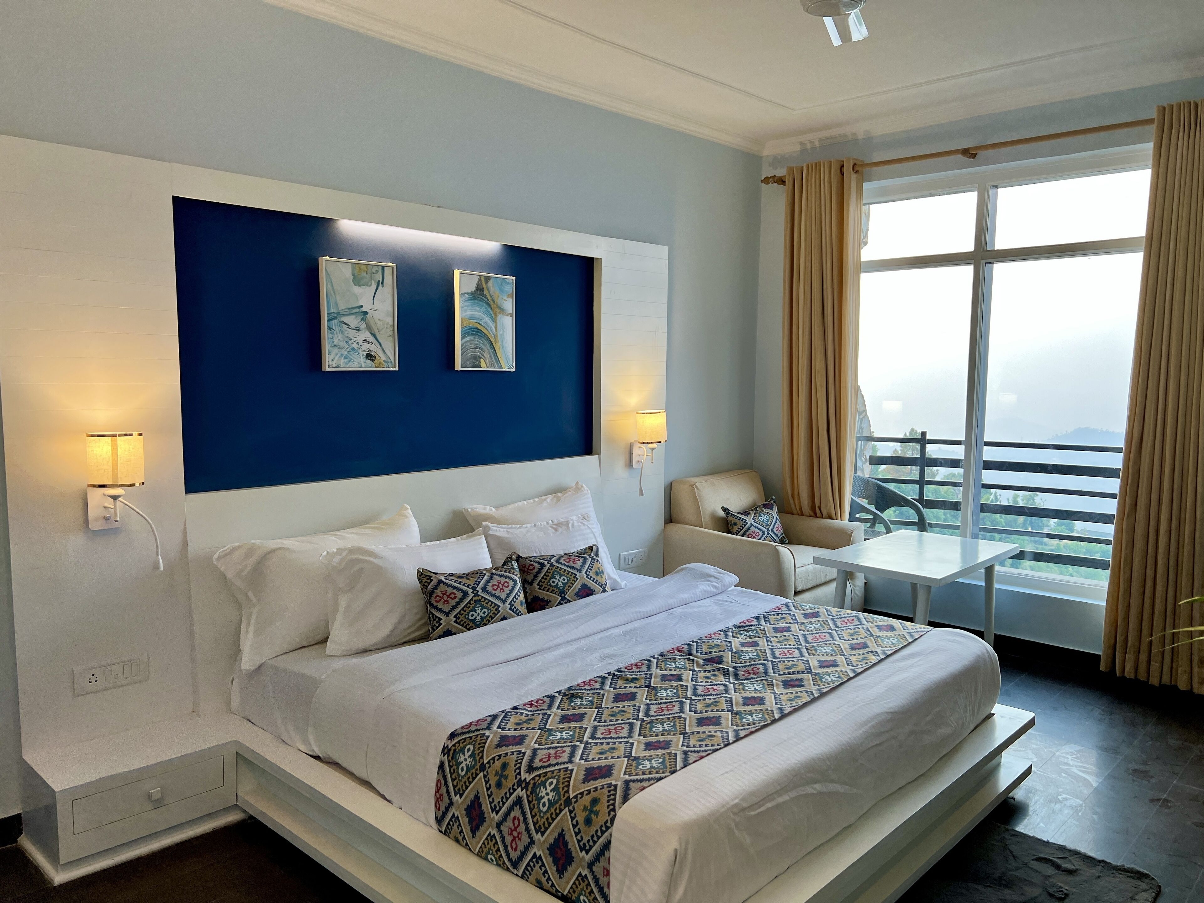A hotel room with a large bed, two wall-mounted lamps, a chair, a balcony with a view, and a window with curtains.