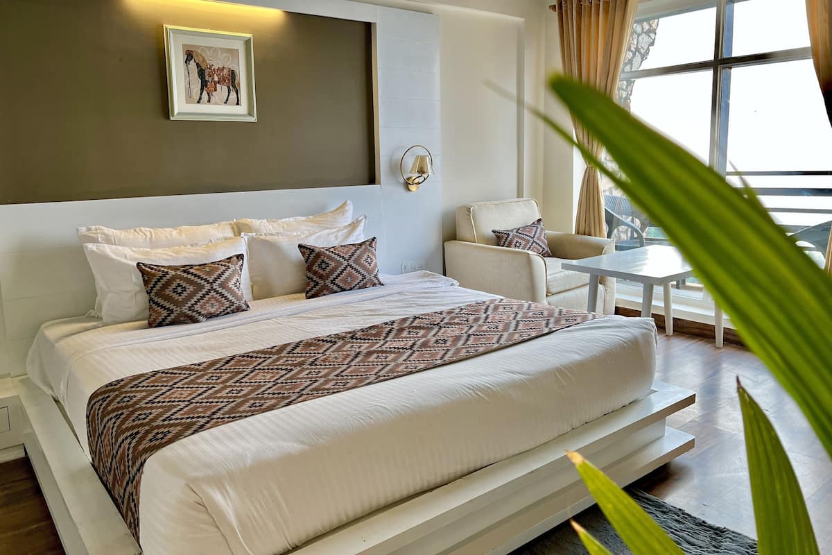 Ein modernes Hotelzimmer mit einem großen Bett, einem kleinen Tisch und einem Sessel. Ein Fenster mit Vorhängen bietet einen Blick nach draußen.