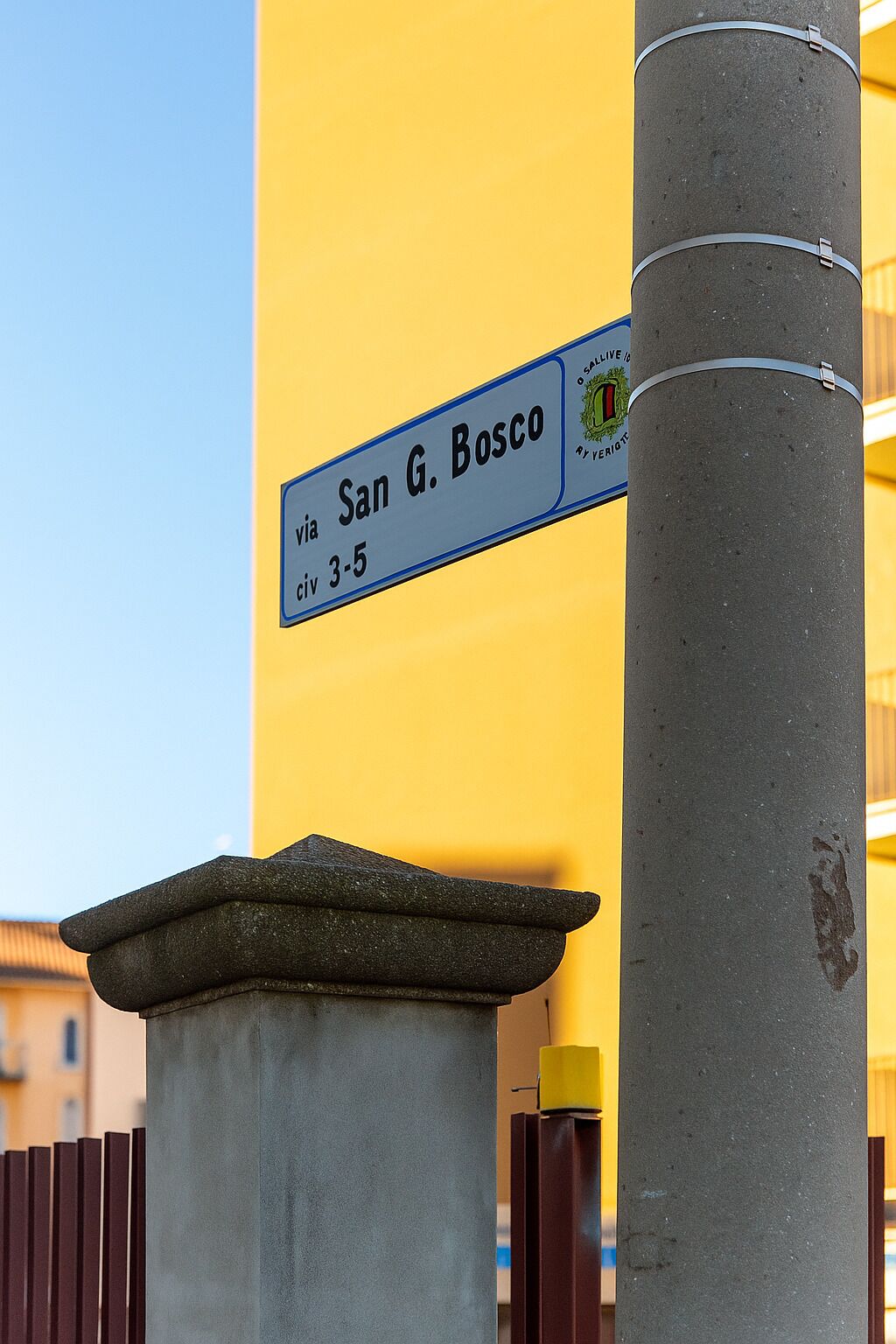 Ein Straßenschild mit der Aufschrift 'San G. Bosco' und 'via 3-5' vor gelbem Hintergrund.