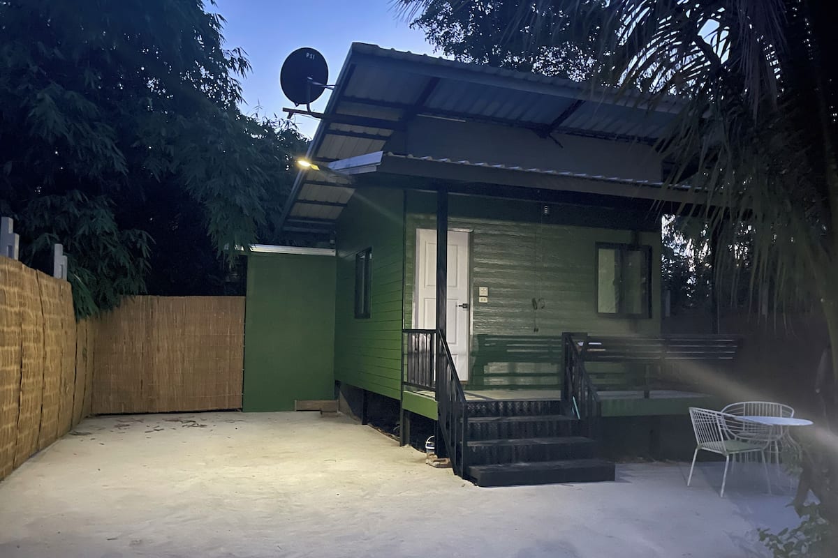 A small, single-story house with a green exterior, a satellite dish, a covered porch, and a small patio area with a chair.