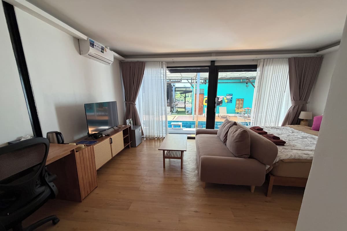 A modern hotel room with a wooden floor, a sofa, a TV, a desk with a chair, and a balcony with a pool.