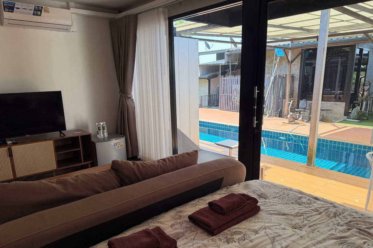 A hotel room with a sofa, a bed with a floral bedspread, a television, a small table with a glass container, and a view of a swimming pool through a glass door.