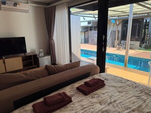 A hotel room with a sofa, a bed with a floral bedspread, a television, a small table with a glass container, and a view of a swimming pool through a glass door. - Morocco blue cafe by chilong river farm  (Kosum Phisai)