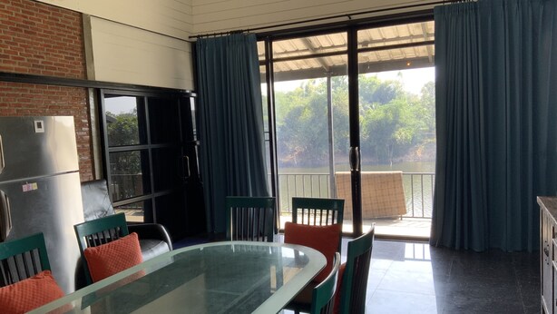 A dining area with a glass table, chairs, a refrigerator, and a view of a lake through large windows. - Morocco blue cafe by chilong river farm  (Kosum Phisai)