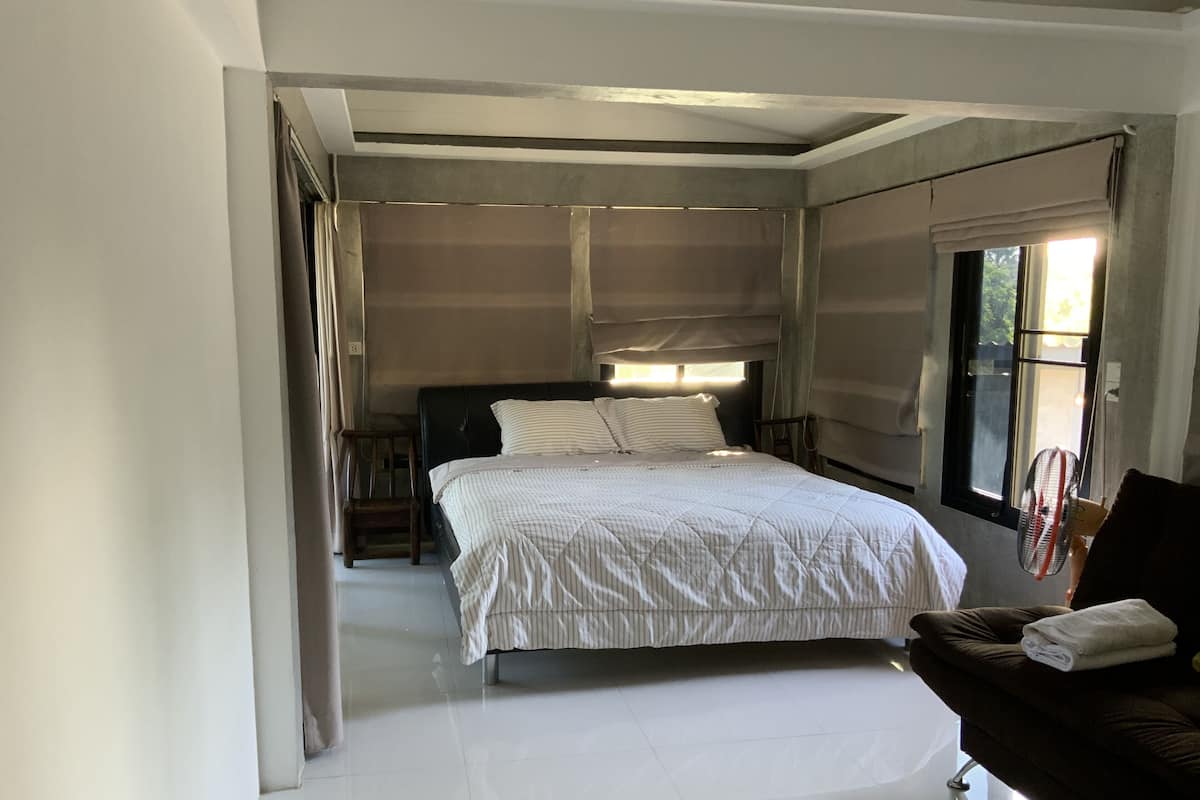 A bedroom with a bed, a nightstand, a chair, a window, and a fan.