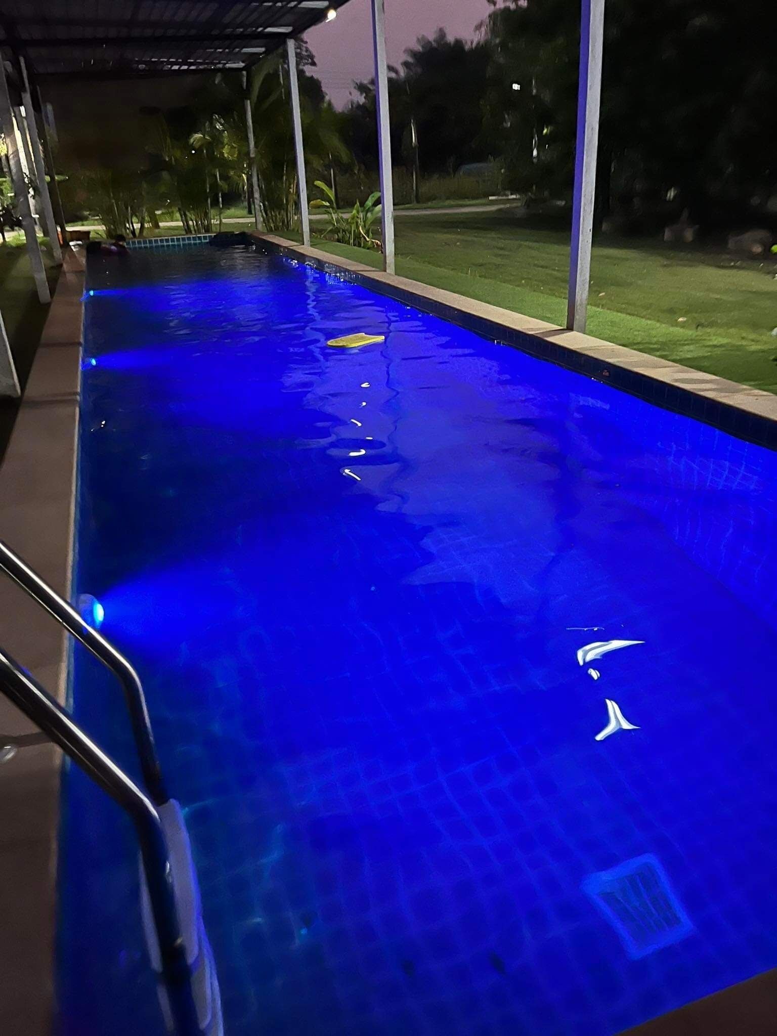 A swimming pool with a blue tint, surrounded by a covered area with a view of a grassy landscape and trees.