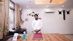 A man standing at a wooden table in a room with a ceiling fan, air conditioning unit, and decorative wall art. - Hotel Lyf Corporate Suites Noida Sec-56 (Noida)