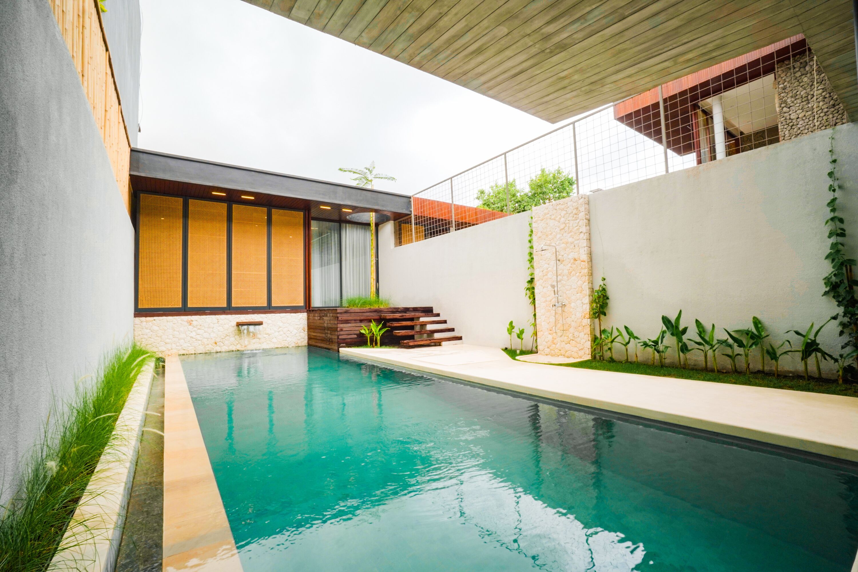A modern pool area with a clear pool, wooden deck, and a stone wall with plants.