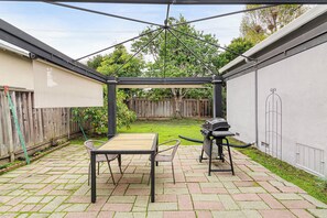 Een overdekt terras met een tafel, stoelen, een barbecue en een houten omheining op de achtergrond.