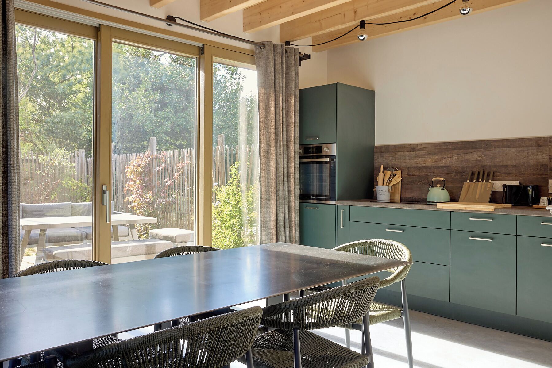 A modern kitchen with green cabinets, a dining area with a table and chairs, and a view of the outdoors through large sliding doors.