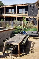 An outdoor dining area with a wooden table and chairs, a cushioned seating area, and a pool in the background.