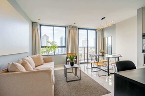 Una sala de estar moderna con un sofá, una mesa de comedor con sillas, una mesita auxiliar con una planta y ventanas grandes con cortinas.