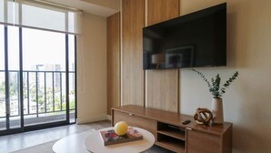 Una sala de estar moderna con un ventanal grande, un televisor de pantalla plana montado en la pared, un mueble de madera, una pequeña mesa redonda con un libro y un objeto amarillo, y una planta en maceta.