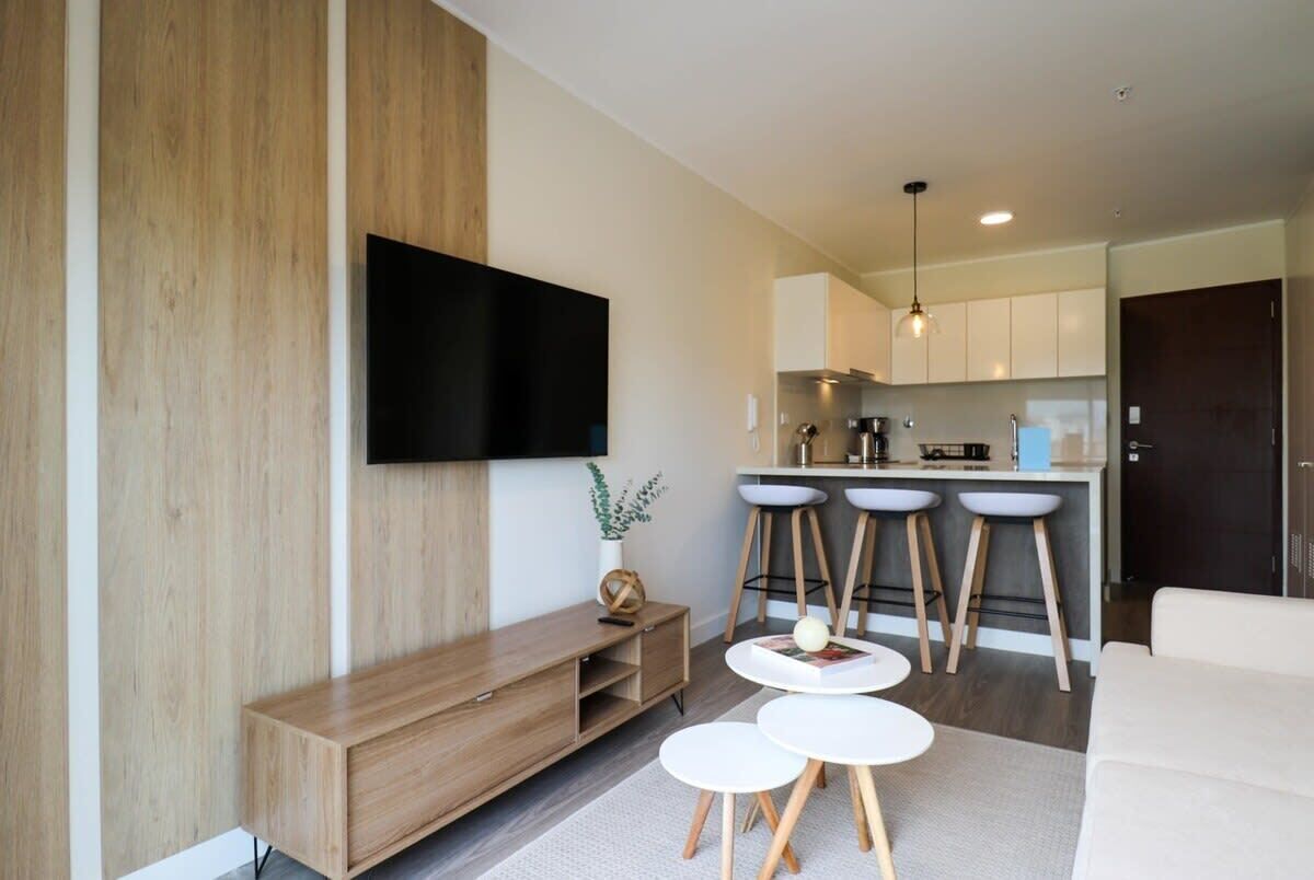 Un salon moderne avec un téléviseur à écran plat fixé au mur, un meuble de rangement en bois, une petite table basse ronde et trois tabourets de bar.