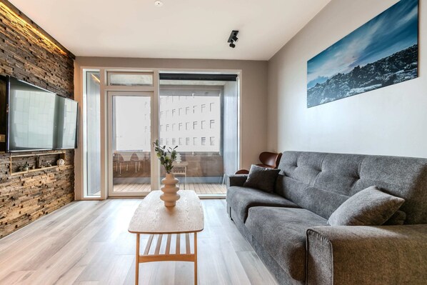 A modern living room with a grey sofa, a wooden coffee table, and a flat-screen TV mounted on a brick wall. - SIF Apartments - 2 bedrooms with 2 bathrooms (Reykjavík)
