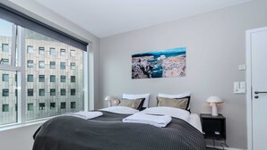 A modern hotel room with a large bed, a nightstand, a lamp, and a painting of a coastal scene.