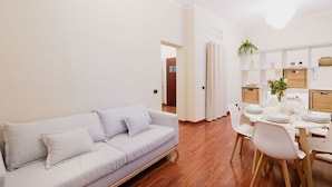 Una sala de estar moderna con un sofá de color claro, un comedor con una mesa blanca dispuesta para cuatro personas, y suelo de madera.