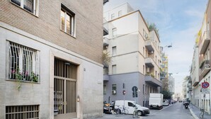 Una calle urbana estrecha con coches y bicicletas estacionados, edificios con balcones y un cielo despejado.