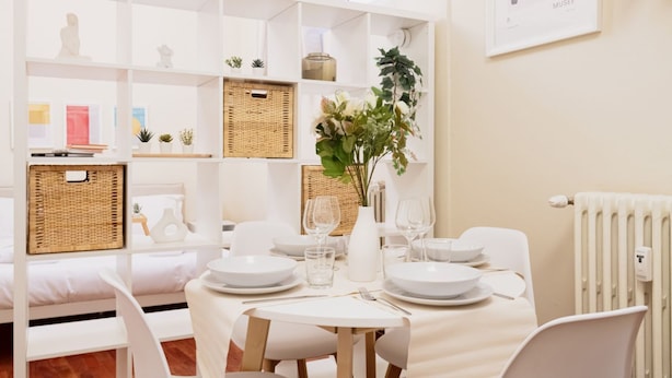 Un comedor con mobiliario blanco, mantel blanco y pared blanca con un cuadro enmarcado.