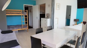 A dorm-style room with bunk beds, a desk, a chair, a dresser, and a kitchenette.