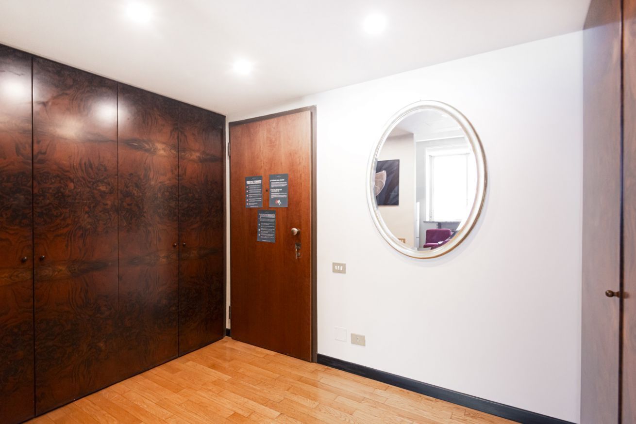 A hallway with wooden flooring, a large wardrobe, a door with a sign, and a round mirror on the wall.