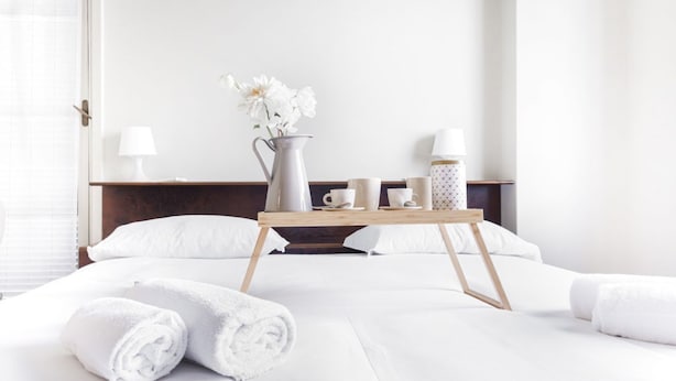 Hotellihuone, jossa on sänky, valkoiset pyyhkeet, yöpöytä, jossa on maljakko täynnä kukkia, sekä tarjotinalusta, jossa on kuppeja ja vesipullo.