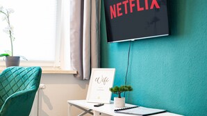 Una camera moderna con una parete verde acqua, una scrivania bianca, una sedia verde acqua, un certificato incorniciato, una piccola pianta in vaso e una TV che mostra il logo di Netflix.