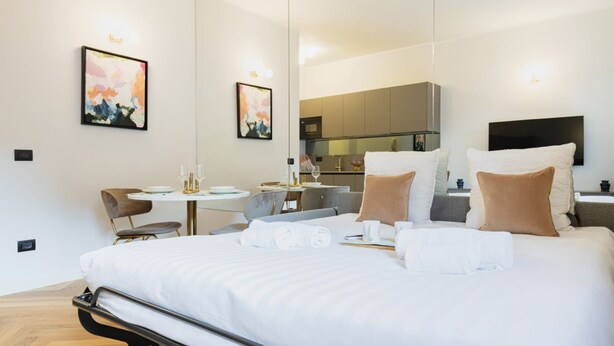 A modern hotel room with a large bed, a small dining table, a chair, a mirror, and a kitchenette in the background. - Easylife - Charming nest in Sant'Ambrogio (Milano)