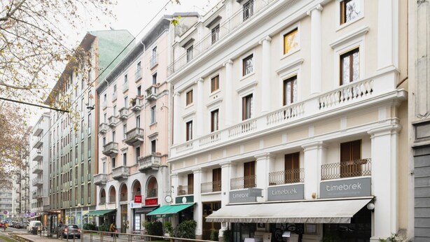 Uma fileira de edifícios com vários andares, com estabelecimentos comerciais no andar térreo, incluindo um café chamado 'timebrek'.