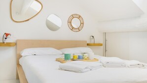 Cama bem arrumada com roupa de cama branca, mesa de cabeceira com abajur e bandeja com xícaras e um livro.