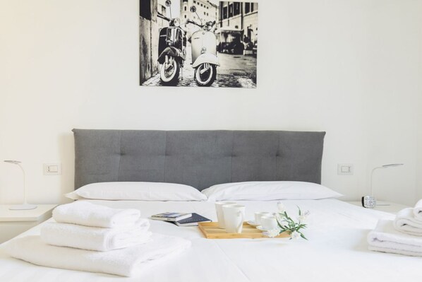 A neatly made bed with white linens, a gray headboard, and a bedside table with a book, a cup, and a tray with a pitcher and flowers.