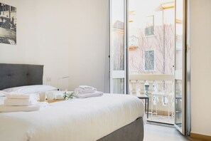 A hotel room with a bed, a small table with towels and a cup, and a view of a balcony with chairs and a railing.