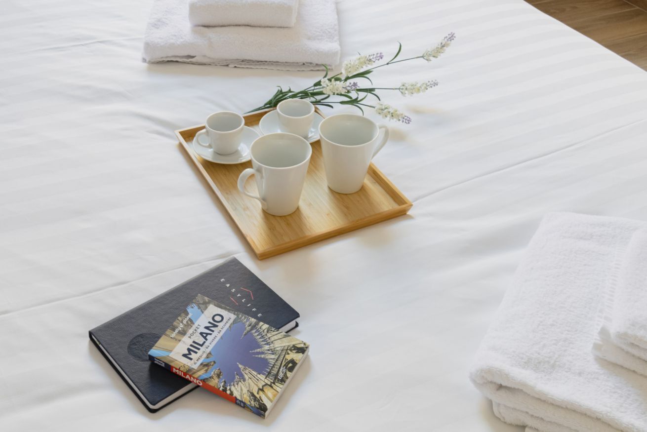 A neatly made bed with white linens, a tray with white cups and saucers, a book titled 'MILANO', and a bouquet of white flowers.