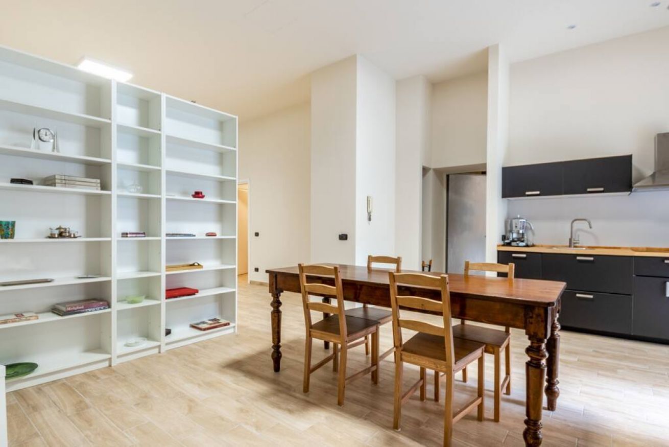 A dining area with a wooden table and chairs, a white bookshelf, and a kitchen with dark cabinets.