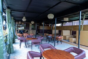 A covered outdoor seating area with tables and chairs, a bar in the background, and a view of the outside. - The Country Inn by Smart Haven (Kigali)